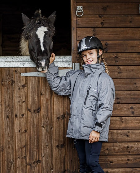 Shortie Child's Oversized Short Waterproof blouson style Riding Jacket Thin Fleece Lining Stowaway Hood in charcoal/penelope pink girl stood with horse 