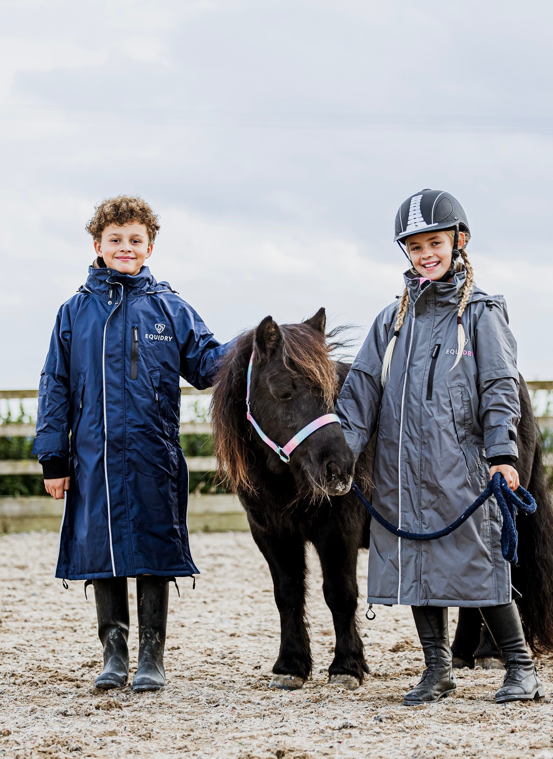 EQUIDRY waterproof horse riding raincoat thin fleece pro ride reflective in navy and charcoal/pale pink children's kids stood in arena with shetland 