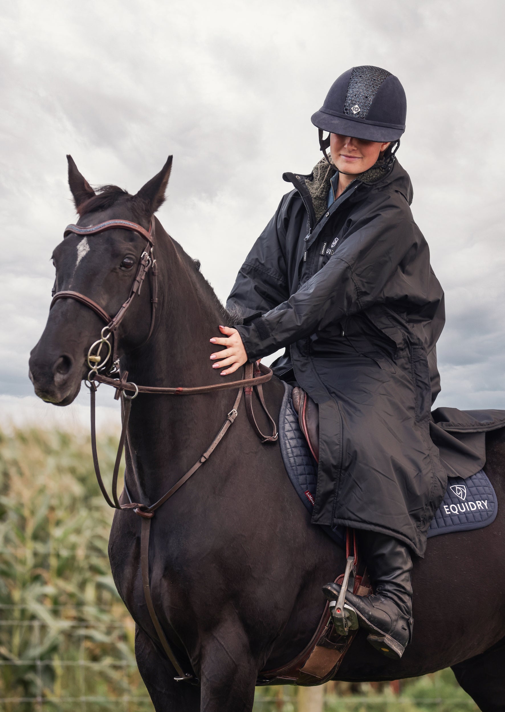 EQUIDRY women's long waterproof horse riding coat in black/olive modelled by rider out hacking, showing storm skirts and side zips